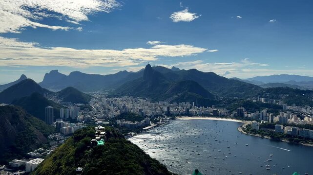 wide shot from Pao de Acucar summit overlooking Botafogo and Flamengo beaches. Features the Rio de Janeiro skyline, Corcovado mountain, and yachts in the Atlantic Ocean under a bright sky.