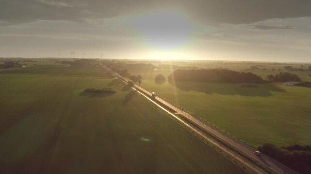 A wide aerial view shows the show truck on a straight country road cutting through green fields. The warm backlight from the setting sun creates a stunning golden atmosphere.