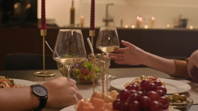 Handheld shot of affectionate Caucasian man and woman drinking white wine, clinking glasses and talking while sitting at festive table with candles during home celebration