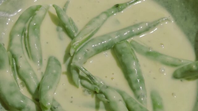 Green chillies are fully coated in a thick, yellow besan (chickpea flour) batter, a common step in the preparation of spicy Indian fritters or pakoras before they are deep-fried to crispy perfection. 