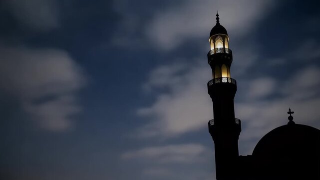 Silhouette of a mosque minaret against a cloudy night sky with illuminated tower