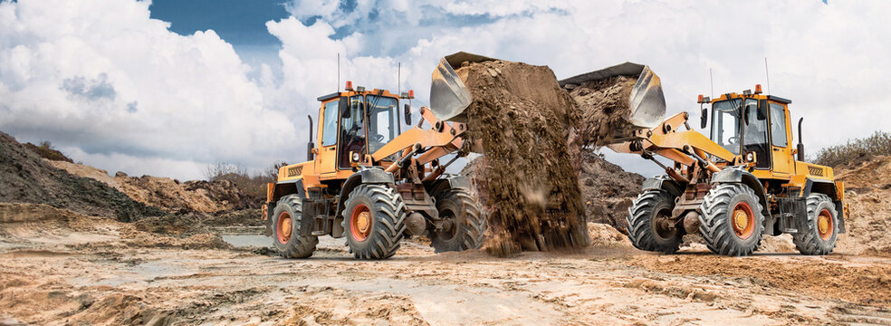 Two loaders move dirt on cracked earth at a construction site with mounds and a cloudy sky above, showing active equipment use in the area