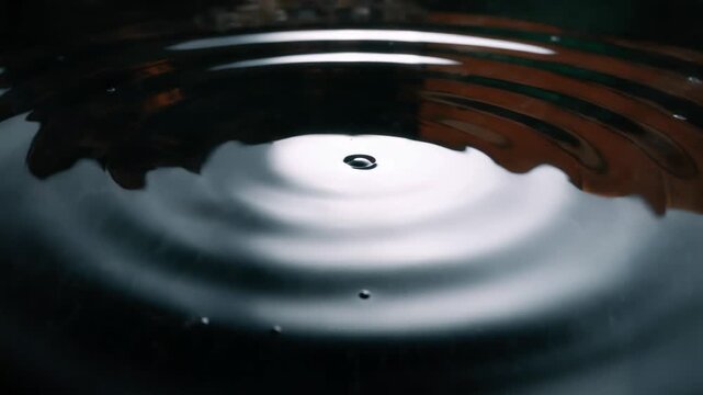 Single drops of dark water fall onto a mirror-like surface, forming even concentric circles and smooth diverging waves