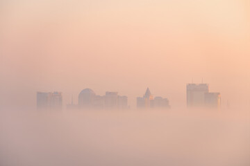 Obraz premium Ethereal cityscape shrouded in dense, atmospheric fog at sunrise or sunset, with prominent buildings softly emerging from the warm, peachy-orange mist. A tranquil and mysterious urban scene.