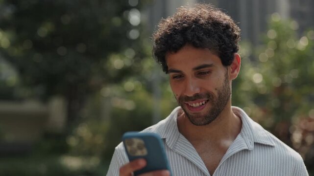 Portrait of positive Hispanic man writes message on smartphone as stands in green urban park slow motion. Young Latin American male scrolls social network on mobile phone in city