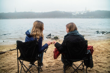 Fototapeta premium Two women sitting in folding chairs on a sandy beach, drinking hot beverages on a foggy day in Kyiv