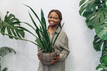 A cheerful African American woman happily holds a large potted plant surrounded by lush, vibrant greenery and a tranquil setting