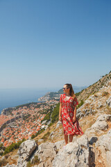 A woman wearing a bright and vibrant red dress stands atop a rocky mountain, gazing out at a stunning coastal view