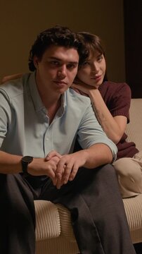 Vertical portrait of happy young Caucasian boyfriend and girlfriend sitting on home sofa, looking at camera while enjoying time together