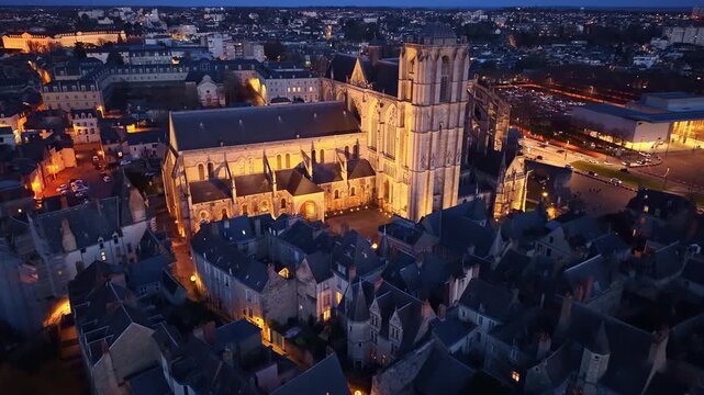 High-angle aerial view of illuminated Cath&eacute;drale Saint-Julien du Mans at night, with the glowing city lights of Le Mans in background, France. Aerial drone forward