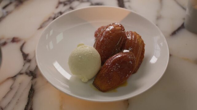A professional chef's hand carefully adds a perfect scoop of white ice cream to a plate featuring three golden, glazed madeleines on a marble surface.