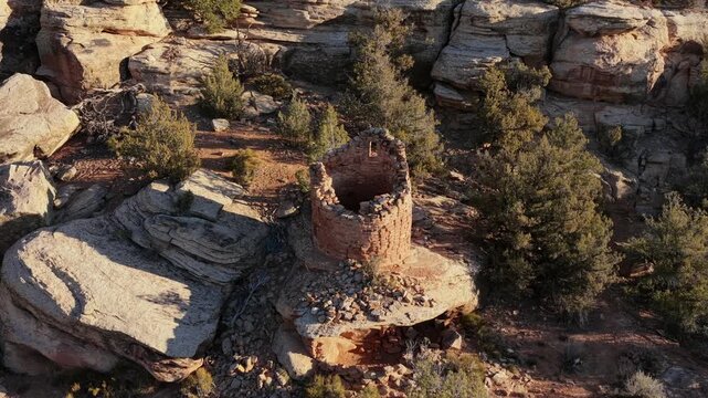 An ancient dry-stone tower sits perched on a rocky mesa above a deep desert canyon. The intricate masonry is surrounded by high-desert vegetation and rugged geological formations.