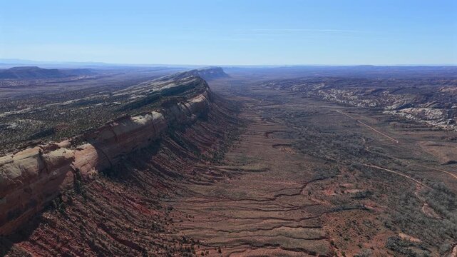 A wide drone view follows a long stone cliff stretching across a dry desert land. The high view shows deep rock layers and a large valley floor under a clear blue sky in the western wilderness.