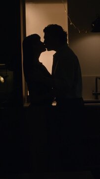 Vertical shot of silhouettes of affectionate man and woman kissing each other closely in dimly lit kitchen