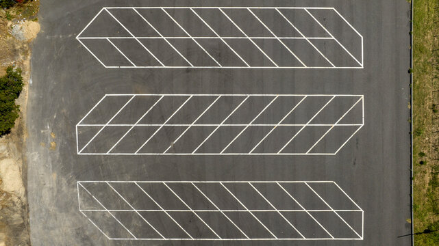 Top-down aerial view of empty parking lot spaces, showing three parallel rows of white diagonal line markings on a dark grey asphalt surface. Zenithal shot of car park.