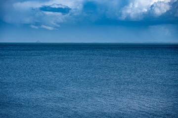 Paysage côtier avec vue lointaine sur le Mont‑Saint‑Michel à travers une légère brume marine, sous un ciel bleu parcouru de rayons lumineux. Végétation en premier plan et vaste étendue d’eau. © d70 Philippe Dougoud
