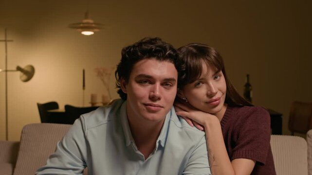 Medium close up portrait of young happy woman leaning on shoulder of Caucasian boyfriend looking at camera in living room under warm lighting