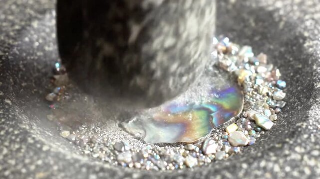 A close-up of a black object crushing an iridescent shell amidst small white stones