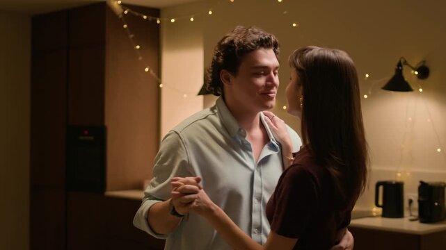 Medium shot of young affectionate couple dancing with passion and looking at each other in cozy home kitchen under warm lighting