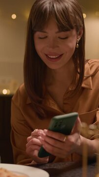 Vertical shot of cheerful Caucasian woman and man laughing while using smartphone sitting at table with wine glasses and homemade dishes