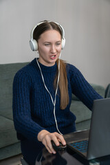 Young woman wearing headphones, engaging in a video call while working from her home office. Focused on her laptop, she talks and navigates with a computer mouse, embodying modern remote work