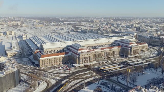 A vast aerial view of Leipzig's snow-covered main station. The historic terminal building and massive platform halls are blanketed in white under bright daylight.