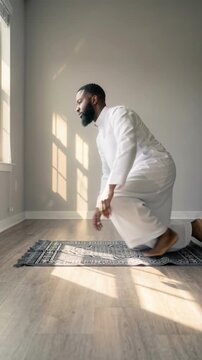 African American Muslim man in white thobe performing salah prayer on a rug at home. Male standing up from prostration in a sunlit room during Ramadan. Islamic faith concept