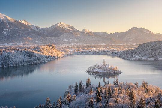Aerial view of the snow-dusted island church amidst the serene lake, framed by majestic, snow-capped mountains, Bled, Radovljica, Slovenia.