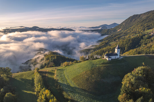 Aerial view of a church standing resolute on a verdant hill, amidst a sea of clouds and rolling green hills, Zgornja Besnica, Kranj, Slovenia.