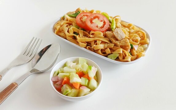 Top view of Asian stir fried flat noodles with vegetables served on a white square plate. Simple minimalist food presentation with fork and spoon on the side, isolated on white background.kwe tiauw