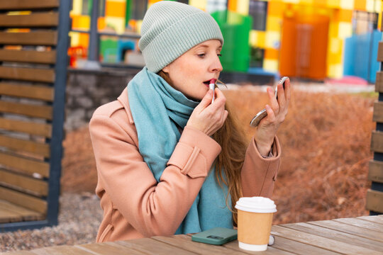 A beautiful woman sits in the courtyard of a house and applies lipstick. It's a cold day outside