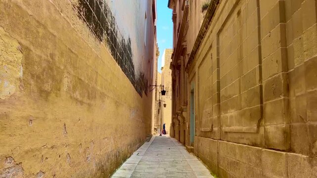 First-person view of walking through the silent city of Mdina in Malta. Ancient yellow stone walls, narrow medieval alleyway under a blue sky, sunny day.