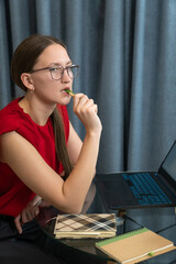 Businesswoman in a red blouse sits at a desk, contemplating work on her laptop. Surrounded by notebooks and books, she holds a pen, suggesting deep concentration and planning. Vertical photo
