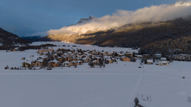 Aerial view of the village nestled in the snow-covered valley, with mountains rising behind under a dramatic sky, Sils im Engadin/Segl, Grigioni, Switzerland.