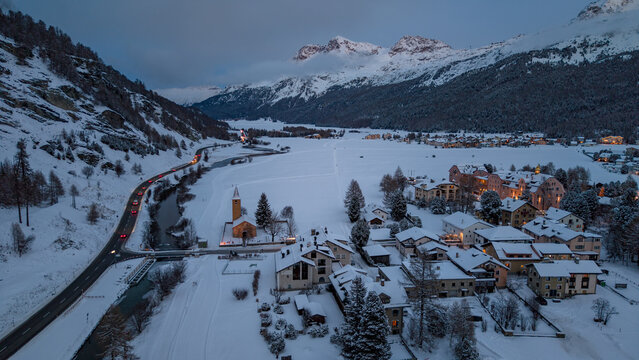 Aerial view of snow-covered village, with a church steeple piercing the twilight sky, nestled at the foot of majestic, snow-dusted mountains, Sils im Engadin/Segl, Grigioni, Switzerland.