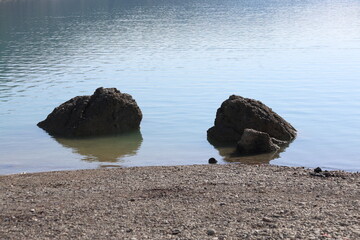 Two Large Rocks Resting in Calm Blue Water by the Shore: Scenic View of a Peaceful Lake with a Pebble Beach Under Bright Daylight