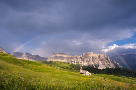 View of a vivid rainbow arcs over a serene meadow, contrasting with the rugged, sunlit Dolomites and a quaint church, Urtijei, Trentino-South Tyrol, Italy.