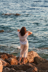 Girl standing on the rocky beach at sunset, looking at the sea and enjoying summer vacation.