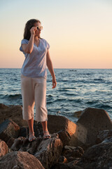 A young woman in sunglasses stands on a rocky beach at golden hour, enjoying the beautiful sunset view and the freedom of a summer holiday by the Mediterranean sea.