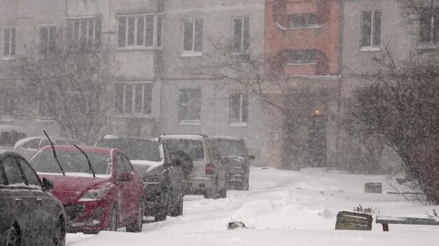 snowfall obscuring parked cars along residential street, flakes drifting past building facades and balconies, muted color palette and accumulating mounds, quiet winter atmosphere with reduced