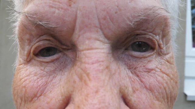 Portrait of mature woman screwing up her gray eyes and then open. Close up wrinkled face of old grandmother looking into camera with a sad sight. Sorrow facial expression of senior lady. Slow motion