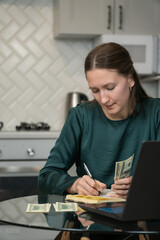 Focused young woman managing home finances, counting cash and using laptop, sitting at glass table in kitchen, managing budget and online banking. Vertical photo