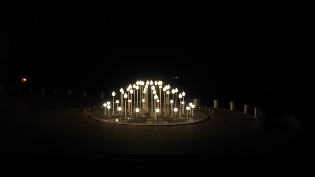Dozens of vintage style white street lamps with glowing spherical globes arranged in concentric circles on dark paved plaza within metal fence at night.