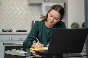 Focused young female freelancer calculating income and expenses, managing finances, counting cash money and making notes in notebook while working remotely at home