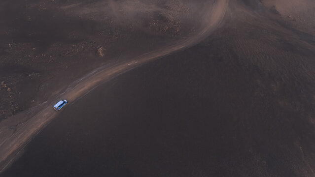 Aerial view of a lone vehicle traversing a stark, dark, volcanic landscape, its blue contrasting vividly against the somber earth tones, Selfoss, Iceland.