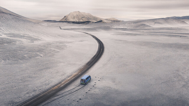 Aerial view of a lone bus winding through the snow-covered landscape, shadowed by distant mountains under a pale sky, Selfoss, Iceland.