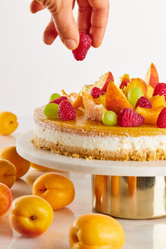 A hand placing a red raspberry on a cheesecake with fruit jelly, on a dessert stand, with apricots on a marble table