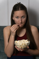 Young woman is sitting on a gray sofa, eating popcorn from a glass bowl and watching a captivating movie on television in her living room. Vertical photo
