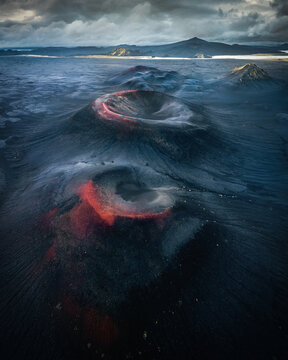 Aerial view of volcanic craters scarred with fiery red deposits against a stark black landscape under a brooding sky, Fjallabak, Iceland.