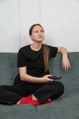 Bored young woman is sitting on a sofa with crossed legs, wearing red socks and a black t-shirt and pants, holding a remote control while watching tv. Vertical photo